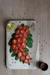 Small red chopped cherry tomatoes lie figuratively on a wooden board with green spinach leaves sprinkled with sesame seeds, a scattering of a mixture of peppercorns, peppercorns in a cup, olive oil, t
