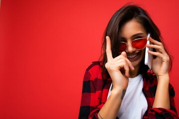 Closeup photo of sexy pretty happy young brunet woman wearing stylish red shirt white t-shirt and red sunglasses isolated over red background communicating on mobile phone looking at camera