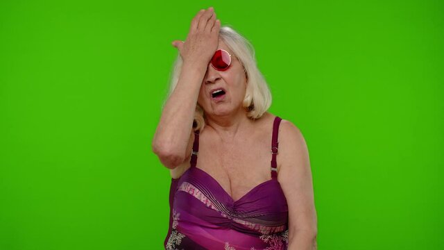Confused senior pensioner woman tourist in swimsuit doing face palm gesture, feeling bored, shame, mistake on chroma key background. Portrait of upset old granny traveler model on summer vacations