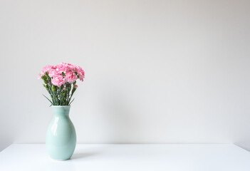 Close up of pink carnations in green vase on whtie table against wall (selective focus)