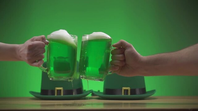two mugs of green frothy beer clink against a background of green Irish hats slow mo