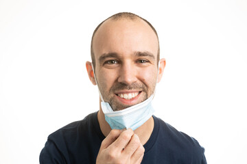 A Caucasian male with a blue t-shirt and stubble facial hair pulling down his blue surgical face mask to smile. White background.