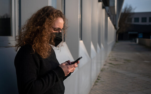 Woman with a mask typing on her smartphone near an office building. Covid 19.