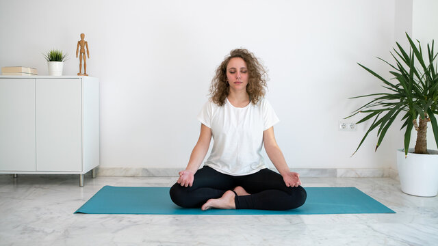 Young woman with closed eyes practicing yoga on a blue mat in the living room.