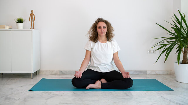 Woman practicing yoga lotus position on a blue mat in her bedroom.
