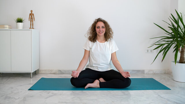 Woman smiling while she does the lotus yoga pose on a blue mat in her home.