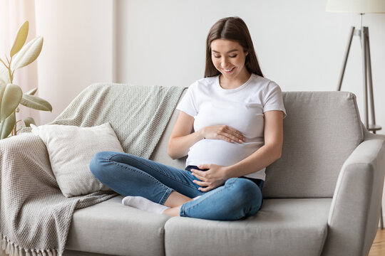 Beautiful young pregnant woman hugging tummy while relaxing on couch at home