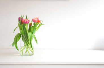 Fresh bright bouquet of colorful Tulips near white wall on wooden shelf in glass vase wit space for text, Spring, April, decoration interion concept modern design. Copy space