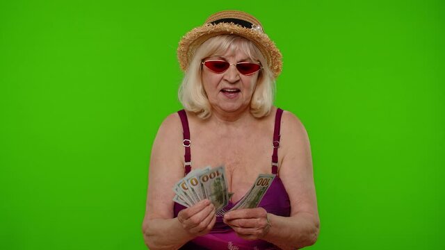 Rich senior woman tourist counting cash enjoying financial independence, waving heap of currency dollar bills on chroma key. Travel, summer holiday vacation. Elderly grandmother in swimsuit. Tourism