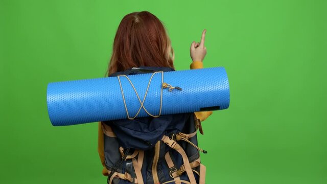 Teenager woman with mountaineering backpack pointing back with the index finger over isolated background chroma key