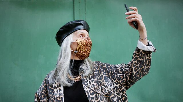 Senior woman with face mask standing outdoors, making selfie. Coronavirus concept.