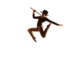 A young female dancer is seen on stage in 
front of a white background. She jumps up 
in mid air posing whilst holding a baton and 
wearing a hat.