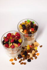 muesli dessert in glass with yogurt and candied or dried fruits with raspberries on top, isolated on white background. raisins, nuts and candied fruits nearby. Granola breakfast