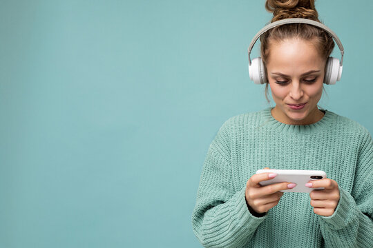 Closeup portrait photo of beautiful positive young woman wearing blue sweater isolated over blue background wearing white bluetooth wireless earphones and listening to music and using mobile phone