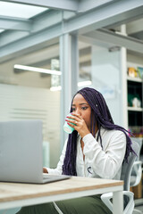 Vertical shot of smiling businesswoman sitting at office desk drinking coffee while working on laptop.