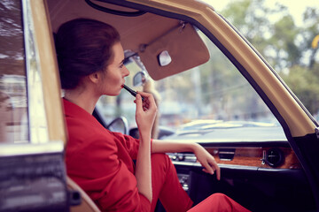 Naklejka premium Beautiful young woman wearing red costume sitting in retro car and paints lips with lipstick.