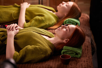 Obraz na płótnie Canvas two redhead ladies lie on bed relaxing after spa treatment massage, in green bathrobes. beauty, healthcare, wellbeing concept