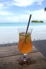 orange cocktail with lemon slices in tulip cocktail glass with straw on brown wooden table with turquoise sea , green island and blue sky as blurred background
