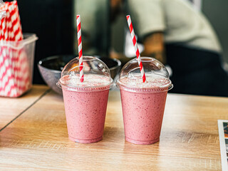 Two pink fruit milk cocktails smoothie in plastic cups with white-red spiral tubes for milkshakes stand on table waiting in cafe