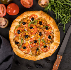 Open pie with red and black tomatoes, olives, arugula and ricotta on paper on a dark background top view