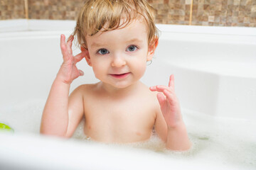 Funny little boy getting a bath in the bathroom. A child bathes in water with soap suds in white bath