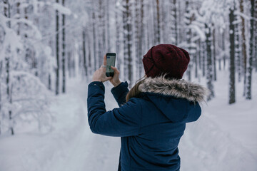 Fototapeta na wymiar Young beautiful happy girl in red winter knitted hat takes photo in winter forest. Travel and active life concept. Outdoors 