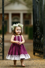 Two year old girl with a flower floral crown and a purple dress outside near an iron gate