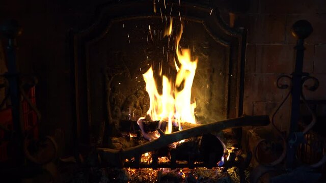 A wood-burning fireplace is lit with wood and flame inside a man throws a piece of wood