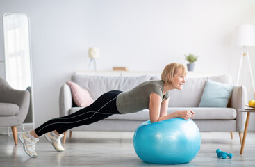 Domestic sports. Athletic mature woman standing in plank on fitness ball, working out core muscles...
