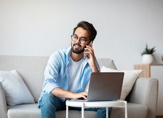 Remote Work. Handsome Arab Freelancer Guy Using Cellphone And Laptop At Home