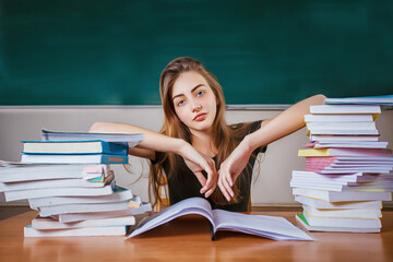 Frustrated female student sitting at the desk with a huge pile of study books in classroom. Young...