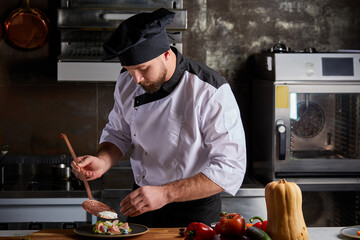 Male cook adding piquancy to dish, finishing with flavouring of meal on plate, wearing cap and...