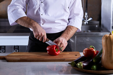 cropped chef man cutting fresh red bell pepper using knife on wooden cutting board. young man in uniform cutting pepper for vegetable salad. concept of healthy eating.