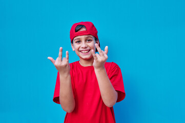 Fototapeta na wymiar Portrait of boy in red t-shirt and cap doing the surfer salute with his hands, on blue background. Copy space