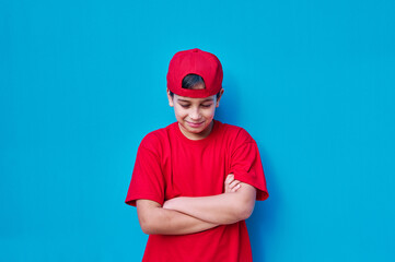Fototapeta premium Portrait of angry boy in red cap and t-shirt with closed eyes and looking down, on blue background. Copy space