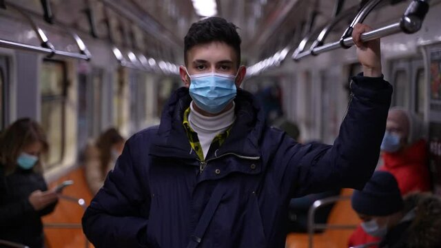 Tall young man wearing medical protective mask and winter jacket standing in metro car holding handrail. Masked teen boy riding in train of underground subway during coronavirus epidemic spread
