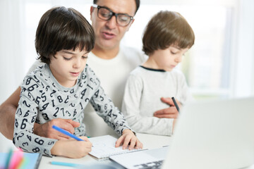Fototapeta premium Teach your child. Cute little latin boy drawing while spending time with his dad and brother at home. Businessman using laptop while working from home and watching children