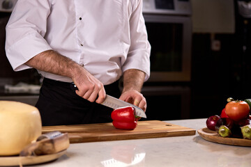 chef cuts red bell peppers on wooden split board to make salad from vegetables, wearing white apron uniform