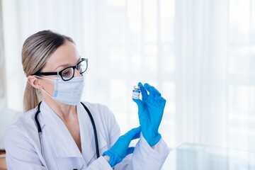 Female doctor in a protective mask and surgical gloves holding an ampoule with vaccine against...