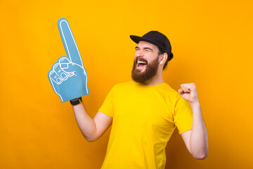 Fototapeta Photo of happy supporter celebrating and pointing with blue fan glove over yellow background. obraz