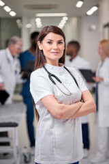 portrait of young caucasian confident doctor woman in white uniform posing at camera in hospital aisle, standing with arms folded. at work. nurse, doctor, medicine concept