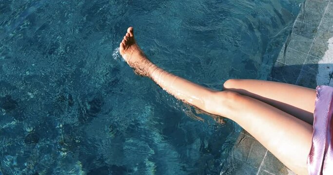 Close-up of the slender legs against the blue water in the resort pool under the sun, in the summer. Woman dangles her legs in the water of the pool. Concept images about, relax, travel, resort. 4K