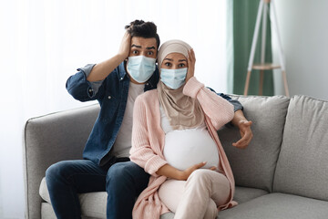 Pregnancy During Pandemic. Shocked Pregnant Muslim Couple In Masks Sitting On Couch