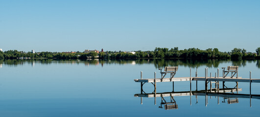 An inviting boat dock is reflected in the glassy water of Lake Irving, the first lake on the Mississippi river, with Bemidji, Minnesota, the 2018 Best Town in Minnesota, seen across the lake.