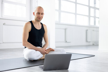 smiling male in sportswear sit on mat with laptop, going to hold online yoga class via webcam,...