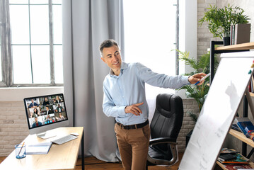 Middle aged successful male top manager or business tutor standing near flipchart in modern office, conducts an online lesson or presentation. Satisfied businessman stylishly dressed on video meeting