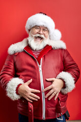joyful senior man in santa hat and red coat laugh at camera holding hands on belly isolated over red studio background. human emotions concept