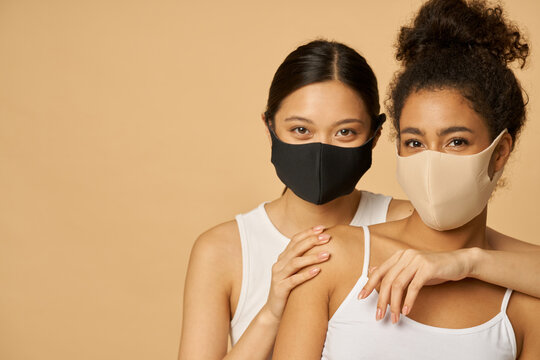 Portrait Of Two Beautiful Young Diverse Women Wearing Protective Facial Masks Looking At Camera While Posing Together Isolated Over Beige Background