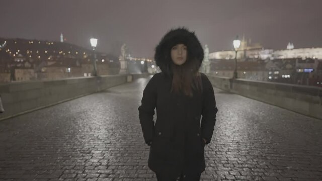 Young woman in jacker with fur hoodie walking on wet pavement of Charles bridge, Prague, Czech Republic on cold winter night