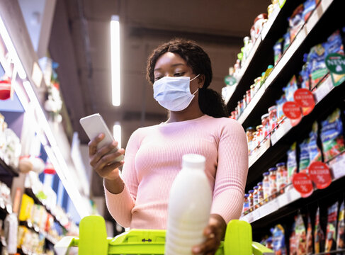 Young Black Woman In Face Mask Holding Smartphone, Going Through Shopping List At Groceries Department Of Supermarket